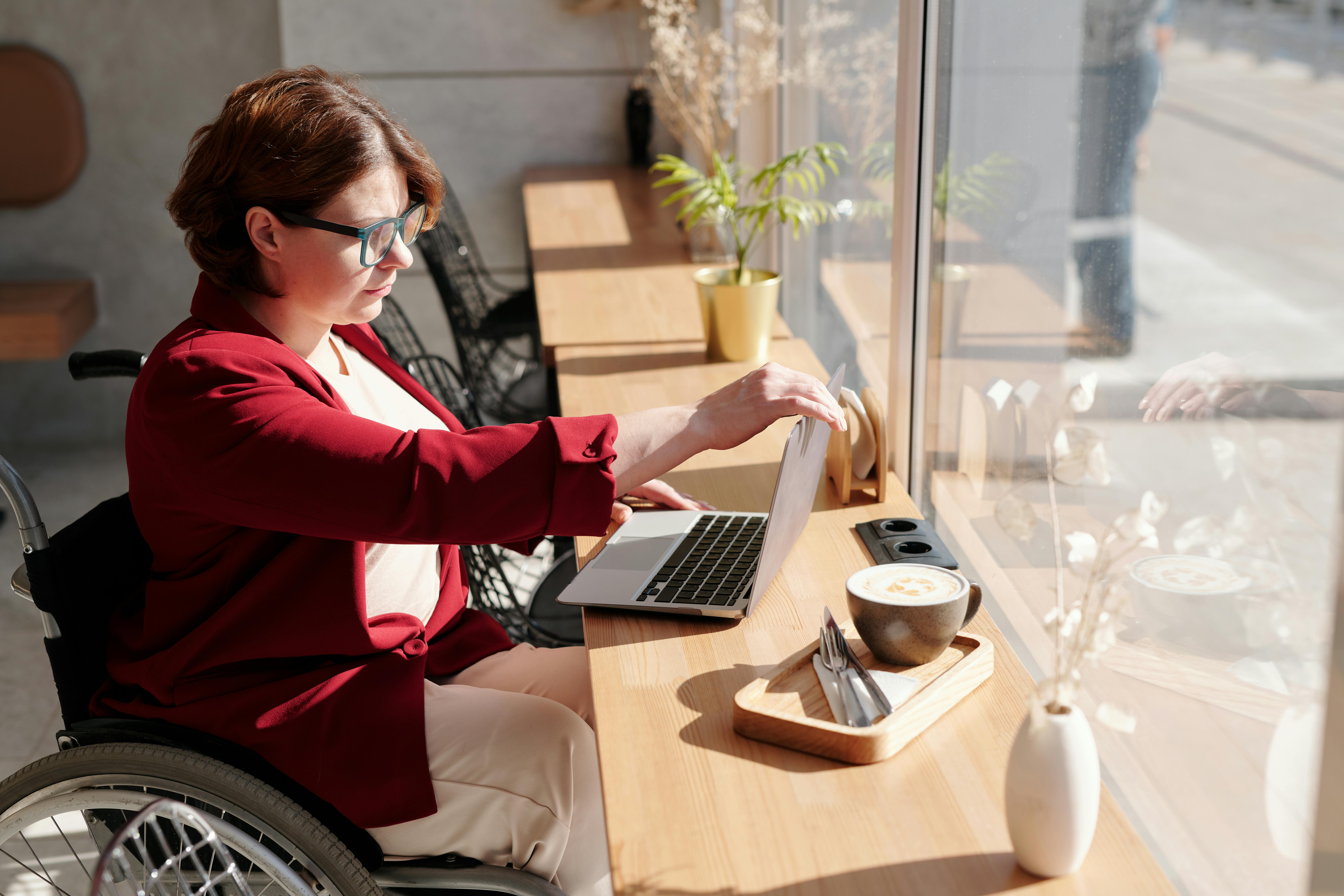 Woman using a wheelchair working on a laptop at a cafe table with coffee, representing independence and inclusive employment.